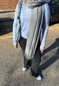 Personne portant un pull bleu clair, un pantalon noir, une écharpe grise et des chaussures bleu clair, debout sur une surface goudronnée ensoleillée près d'une voiture noire.