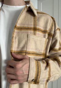 Fleece košile v béžové barvě s žlutým a světle růžovým kostičkovaným vzorem. Vybavena knoflíkovými manžetami a náprsní kapsou s vyšitým logem.