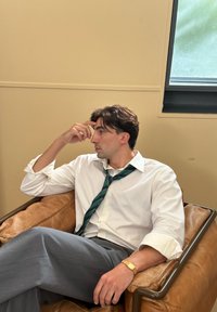 Vīrietis sēž brūnā ādas krēslā, tērpies baltā kreklā, zaļā un tumši zilā svītrā ietērptā kaklasaite un pelēkos biksēs, ar pulksteni uz rokas.