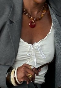 Weißes Top mit gerafftem Vorderteil und Satinschleife, unter einem grauen Blazer getragen. Die Halsketten sind mit Bernsteinen und einem roten runden Anhänger versehen. Goldene Ringe und gestapelte Armreifen.