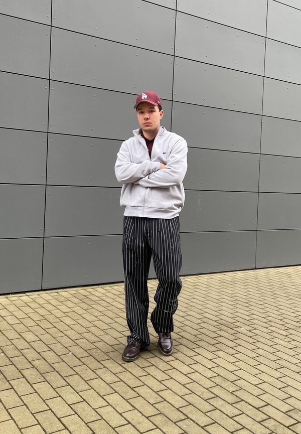 Siv zip-up kapuca, maroon kapa, prugaste hlače crne i bijele boje te tamnosmeđe cipele. Stoji naslonjen na sivi zid na kaldrmi.