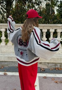 Grijze gebreide oversized trui met rode accenten, grafische patronen en brede mouwen, gecombineerd met rode broek en een rode pet.