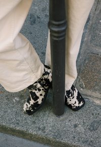 Cizme ankle cu imprimare în alb și negru, cu toc gros, având un detaliu cu cataramă, purtate cu pantaloni de culoare deschisă, poziționate lângă un stâlp metalic.