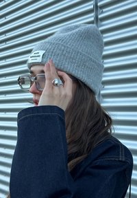 Γκρίζα πλεκτή σκούφια με ετικέτα, διαφανή γυαλιά και ασημένιο δαχτυλίδι φορεμένο σε ένα χέρι. Σκούρο τζιν μπουφάν με εμφανές ραψίμο.