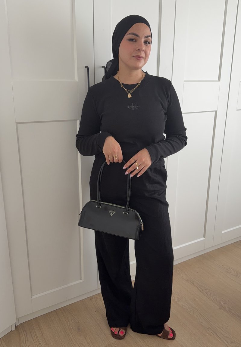 Femme vêtue entièrement de noir avec un foulard, tenant un sac à main noir, debout devant des portes d'armoire blanches sur un sol en bois.