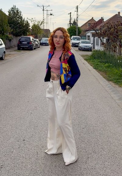 Joven mujer con cabello rizado rojo, chaqueta colorida, top corto rosa y pantalones blancos anchos, de pie en una calle residencial con coches aparcados.