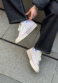 Personne portant des baskets blanches avec un logo étoile, un jean noir à jambes larges, des chaussettes blanches et une veste matelassée, ajustant le bas de son pantalon tout en étant assise sur un pavé en pierre.