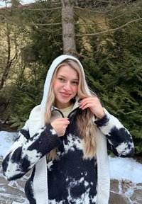 Jeune femme aux longs cheveux blonds fermant sa veste à capuche noire et blanche à motifs, en plein air avec de la neige et des arbres à feuilles persistantes en arrière-plan.