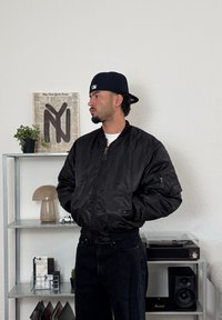 Jachetă bomber neagră cu fermoar frontal și buzunare laterale, purtată peste o cămașă albă. Asortată cu blugi negri, pe un fundal minimalist.