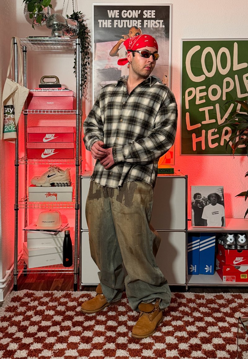 Homme portant un bandana rouge, des lunettes de soleil, une chemise à carreaux et un pantalon ample, debout dans une pièce avec des boîtes de sneakers et des affiches disant "NOUS VERRONS LE FUTUR EN PREMIER" et "LES GENS COOL VIVENT ICI."