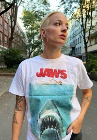 Weißes Baumwoll-T-Shirt mit einem grafischen Druck eines Hais und eines Schwimmers, versehen mit dem Text "JAWS" in fetten roten Buchstaben. Kurze Ärmel, runder Halsausschnitt.