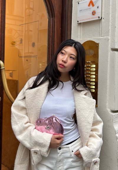 Joven mujer con chaqueta y pantalones blancos sostiene un bolso rosa brillante, de pie junto a una puerta de madera con un panel de intercomunicador dorado en un edificio.