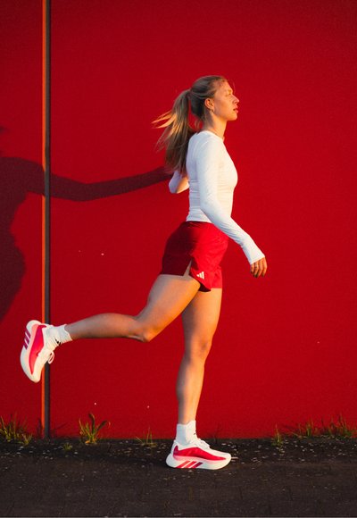 Joven mujer con pantalones cortos rojos y camiseta blanca de manga larga estirando la pierna derecha hacia atrás, de pie contra una pared roja brillante al aire libre.