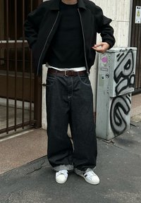 Personne portant un jean large bleu foncé, des baskets blanches, une veste noire et un pull noir, debout près d'un boîtier utilitaire tagué sur un trottoir en ville.