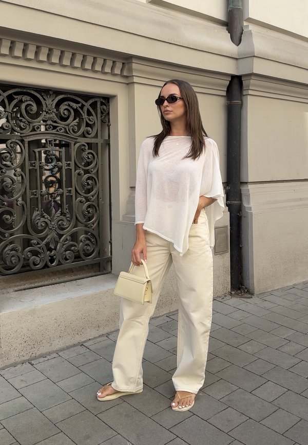 Vrouw met zonnebril, losse witte top, wijde beige broek en sandalen, houdend een kleine beige handtas, staande op een geplaveide stoep nabij een gebouw.