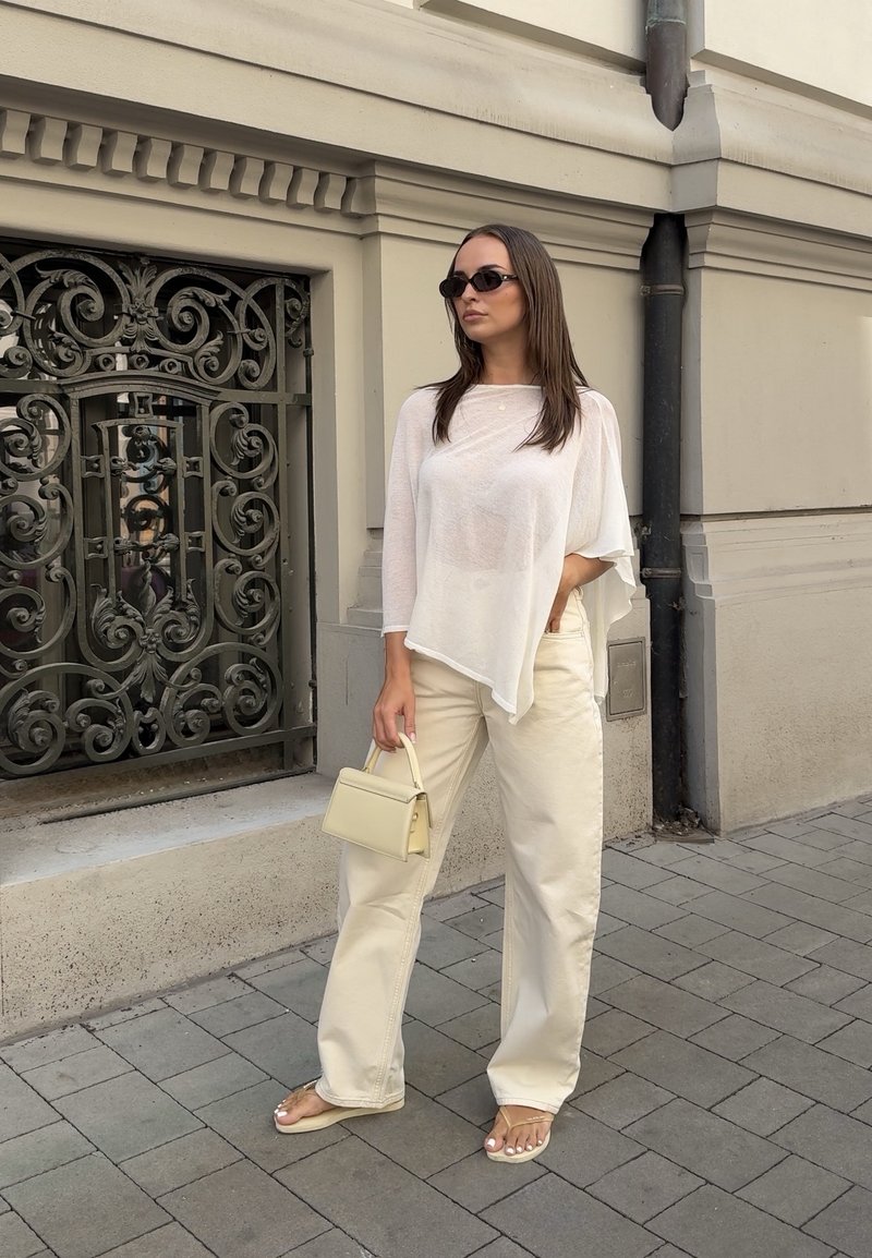 Femme portant des lunettes de soleil, un haut blanc ample, un pantalon beige large et des sandales, tenant un petit sac à main beige, debout sur un trottoir pavé près d'un bâtiment.