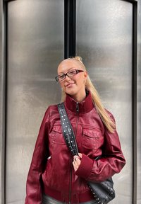 Giacca di pelle rossa con due tasche frontali, colletto alto e polsini a coste. Borsa a spalla nera decorata con borchie argentate. Texture liscia.