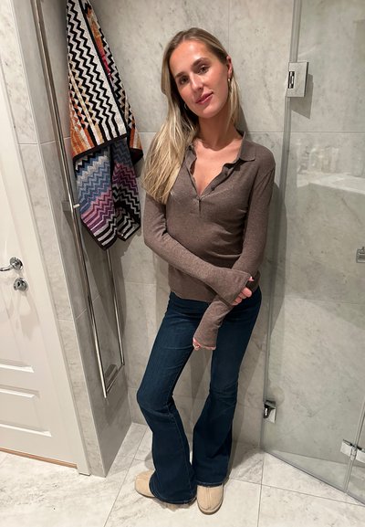 Camisa marrón de manga larga con cuello, jeans acampanados azules, zapatillas de color crema, toalla estampada colgada detrás en una pared de azulejos.