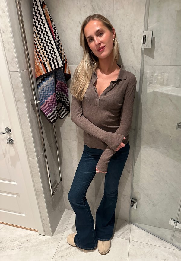 Camisa marrón de manga larga con cuello, jeans acampanados azules, zapatillas de color crema, toalla estampada colgada detrás en una pared de azulejos.