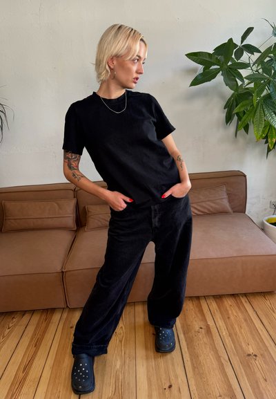 Camiseta de algodón negra combinada con jeans anchos negros. El modelo lleva zuecos negros. Diseño simple, sin patrones ni logotipos visibles.