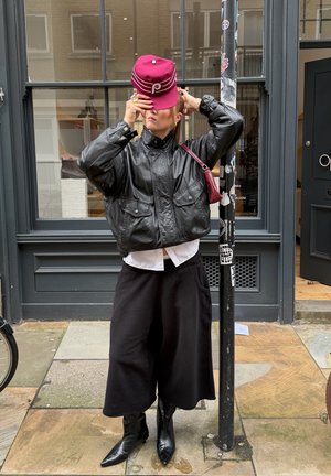 Persona ajustándose una gorra marrón, vistiendo chaqueta de cuero negra, pantalones anchos negros y botas puntiagudas, de pie junto a un poste cubierto de pegatinas en la acera.
