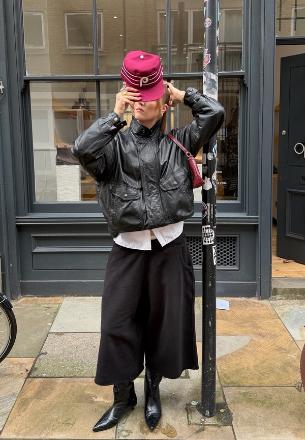 Personne ajustant une casquette bordeaux, portant une veste en cuir noire, un pantalon large noir et des bottes pointues, debout à côté d'un poteau couvert d'autocollants sur le trottoir.
