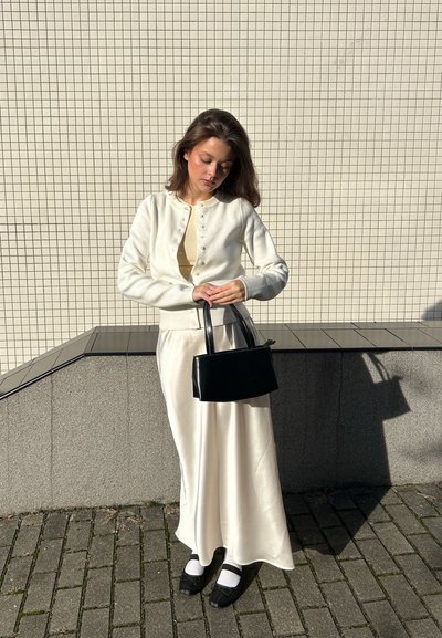 Cárdigan blanco con detalles de botones, blusa beige, falda blanca de satén, bolso negro y zapatos negros con calcetines blancos. Fondo de azulejos texturizados.