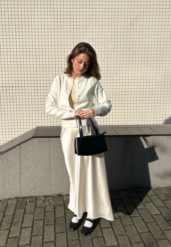Cardigan blanc avec des détails de boutons, haut beige, jupe blanche en satin, sac à main noir et chaussures noires avec des chaussettes blanches. Fond texturé en carreaux.