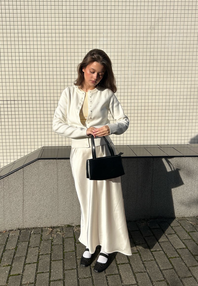 Weißer Cardigan mit Knopfdetails, beiges Oberteil, satinierter weißer Rock, schwarze Handtasche und schwarze Schuhe mit weißen Socken. Strukturierten Hintergrundfliesen.