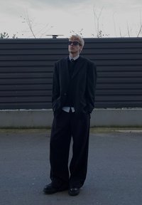 Jeune homme portant un costume noir surdimensionné et des lunettes de soleil se tient dans une rue déserte avec une clôture sombre et des arbres nus en arrière-plan.