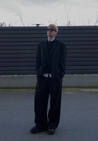 Joven vestido con un traje negro extragrande y gafas de sol, de pie en una calle vacía con una cerca oscura y árboles desnudos en el fondo.