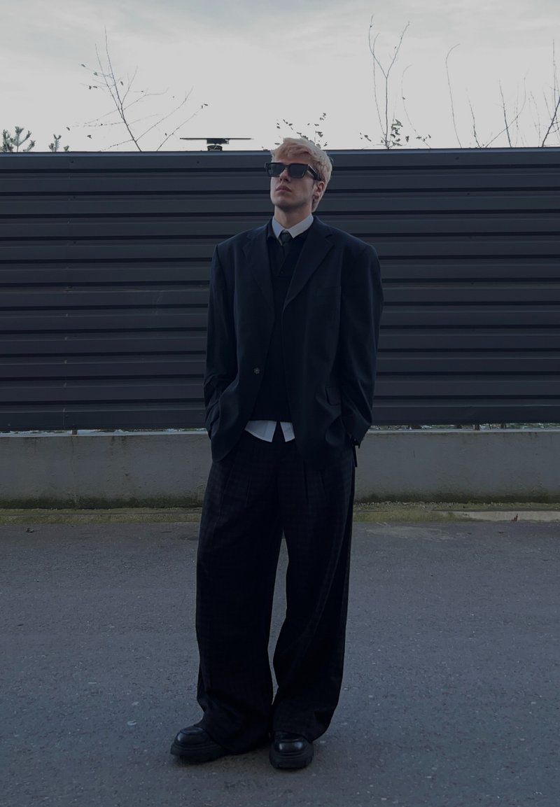 Jeune homme portant un costume noir trop grand et des lunettes de soleil, debout dans une rue vide avec une clôture sombre et des arbres dénudés en arrière-plan.