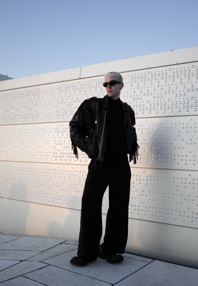 Chaqueta de cuero negra con detalles de flecos, cuello de tortuga negro, pantalones negros holgados y gafas de sol negras, contra una pared blanca texturizada.