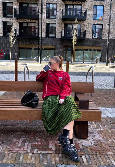 Mujer con gafas de sol y sudadera roja sentada en un banco de madera al aire libre, bebiendo de una taza, con una bolsa negra a su lado.