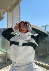 Giacca a vento bianca e verde con design a strisce, polsini elastici e collo alto; indossa cuffie d'argento over-ear.