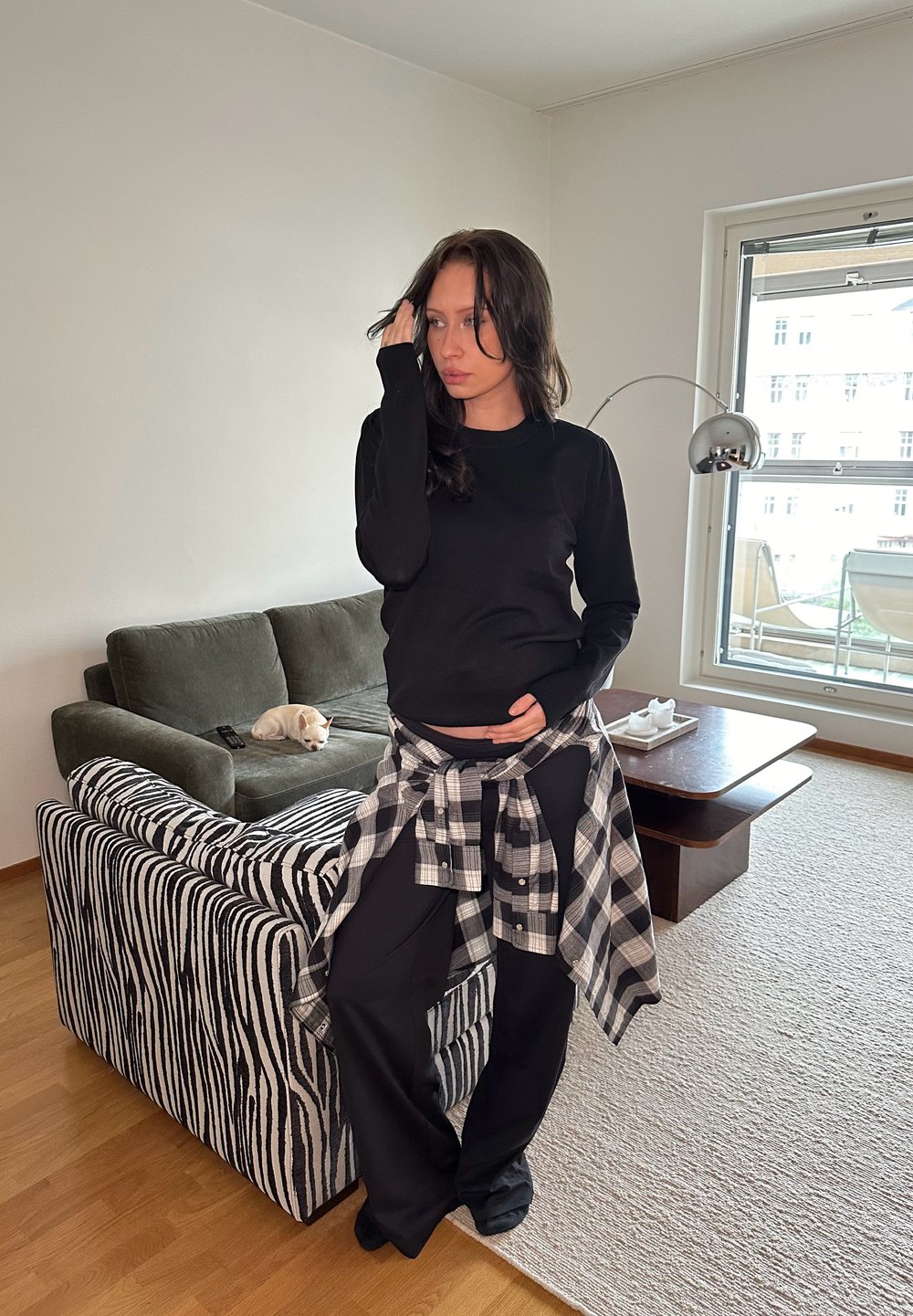 Fekete hosszú ujjú felső, laza fekete nadrág és a derékon megkötött kockás flaneling. Zöld kanapé csíkos mintázattal a háttérben.