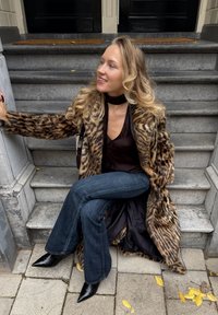 Kvinne med langt blondt hår, iført en leopardmønstret pelsjakke, svart topp, blå jeans og svarte spisse støvler, sittende på betongtrapper utendørs.