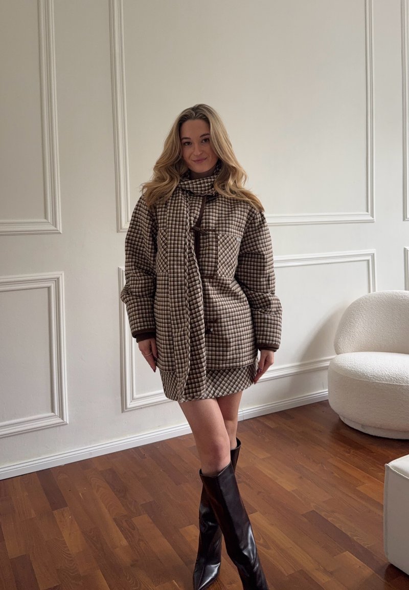 Femme debout à l'intérieur, portant un manteau-robe à carreaux marron et des bottes noires jusqu'aux genoux, avec des murs clairs et un sol en bois en arrière-plan.