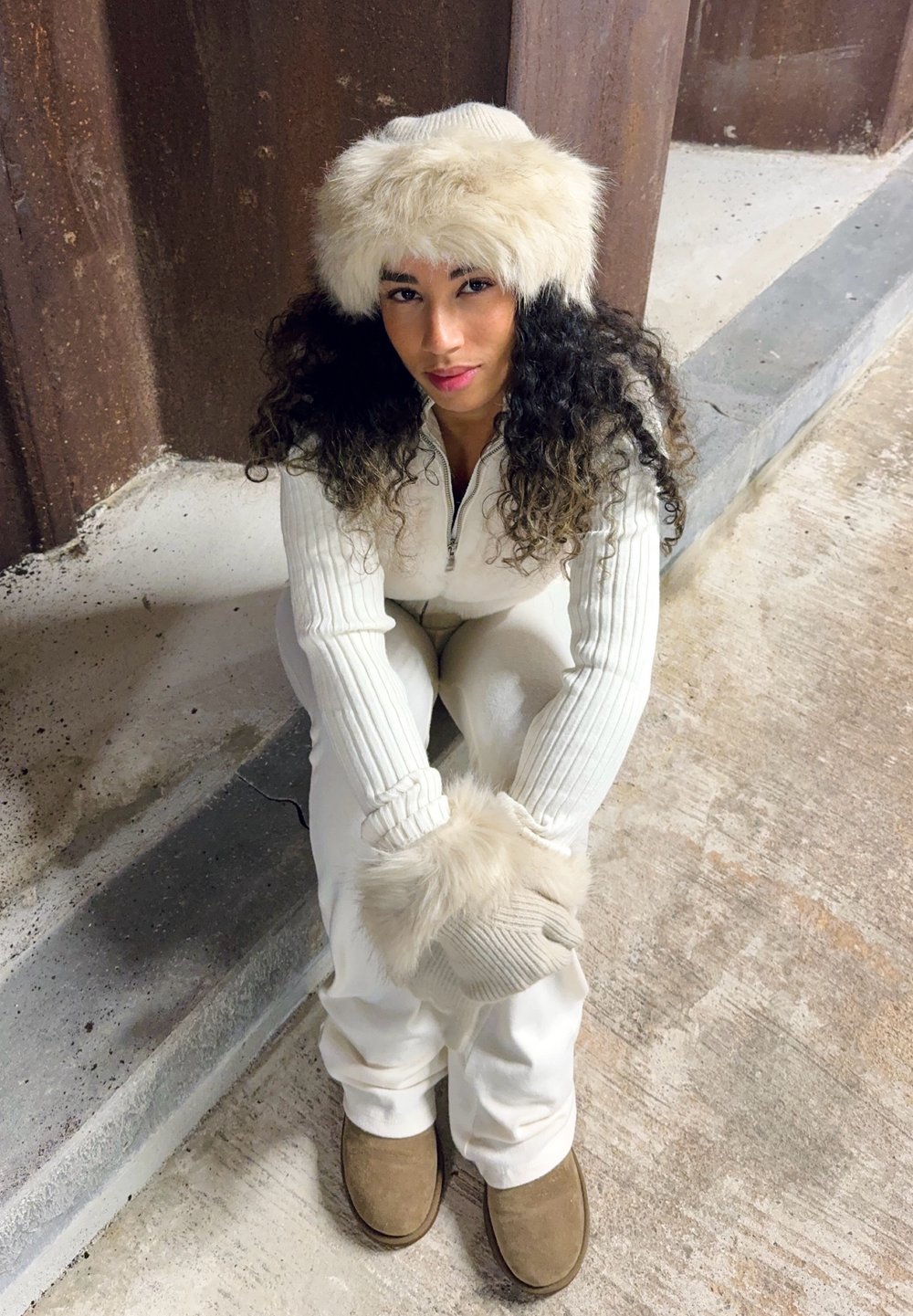 Jumpsuit bianco a costine con zip a un quarto, abbinato a guanti con dettagli in pelliccia e un cappello di pelliccia color crema. Stivali marroni completano il look invernale.