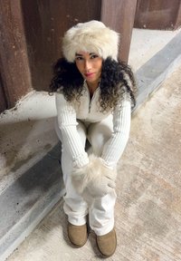 Jumpsuit bianco a costine con zip a un quarto, abbinato a guanti con dettagli in pelliccia e un cappello di pelliccia color crema. Stivali marroni completano il look invernale.