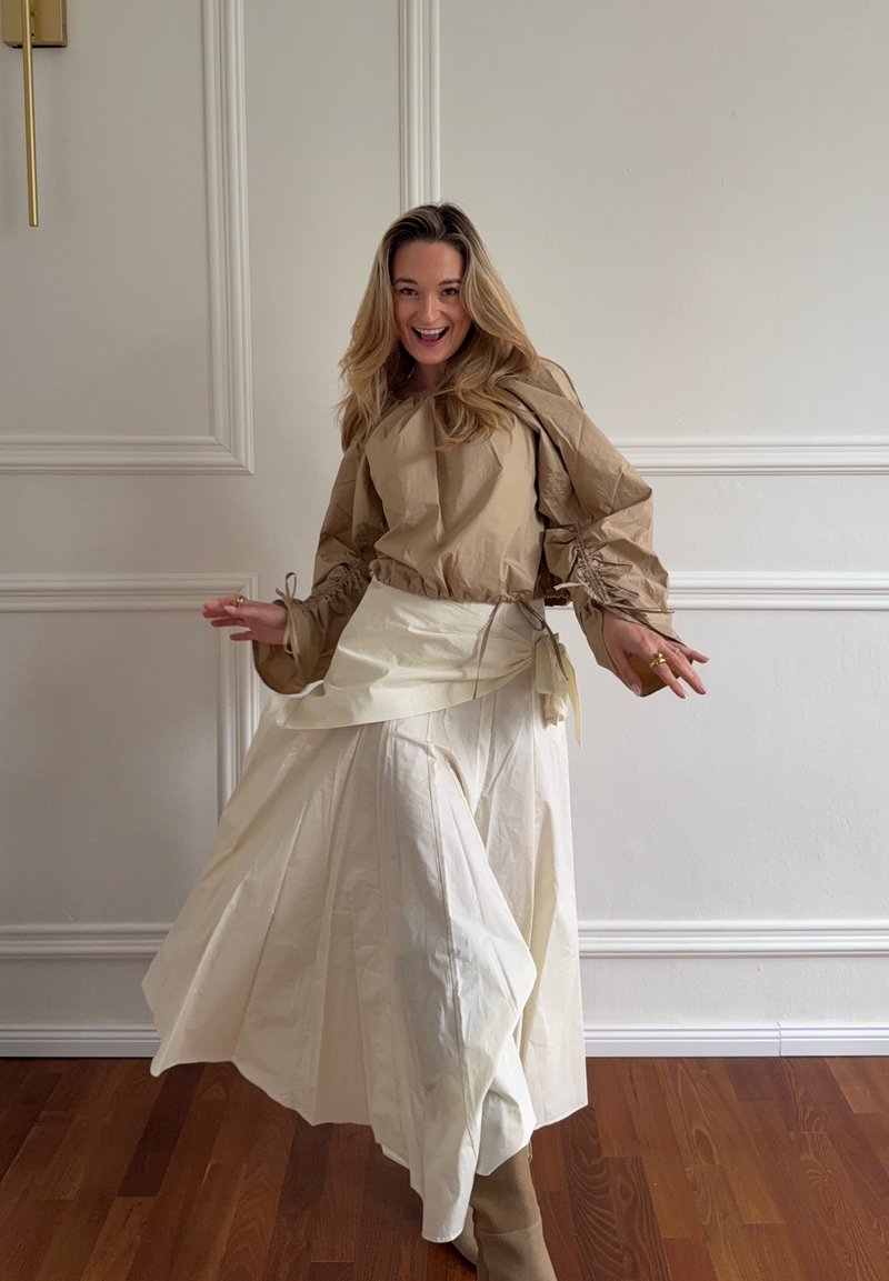 Femme souriante en tournant dans un haut beige à manches bouffantes, une jupe longue crème et des bottes marron clair devant un mur blanc à panneaux et un sol en bois.
