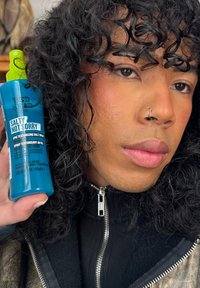 Una bottiglia spray blu etichettata "Salty Not Sorry" con un tappo verde, tenuta accanto a una persona con capelli neri ricci, che indossa una giacca nera.