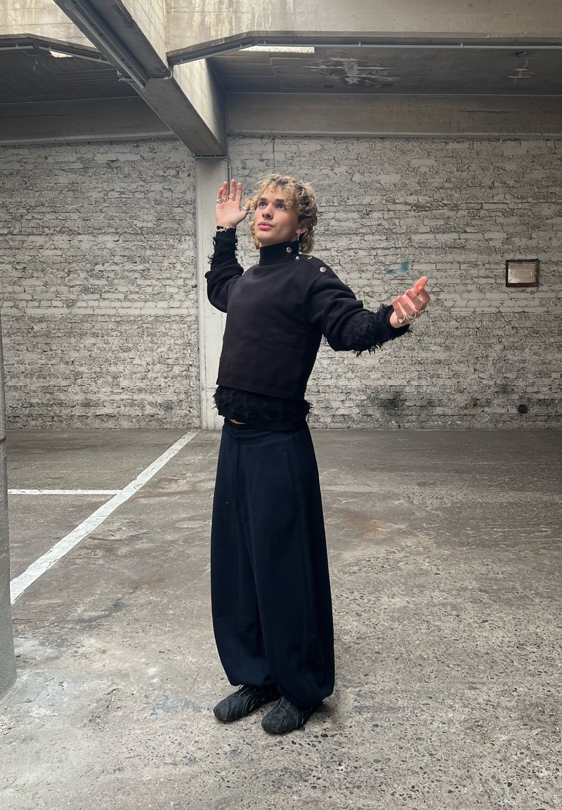 Black cropped jumper with long, textured sleeves and button detail, paired with loose, navy wide-leg trousers; stands in an industrial setting.