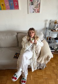 Peignoir blanc douillet avec broderie colorée, pantalon de pyjama blanc et chaussons pelucheux avec motif cerise. Tenant une tasse blanche, assise sur un canapé beige.