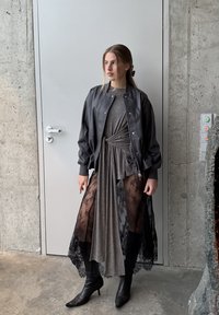 Blouson aviateur noir par-dessus une robe grise ; la robe présente des panneaux en dentelle transparente, des bottes noires montant jusqu'au genou. Textures lisses, design superposé.