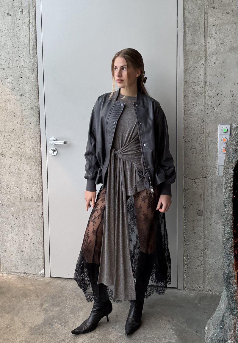 Blouson aviateur noir par-dessus une robe grise ; la robe présente des panneaux en dentelle transparente, des bottes noires montant jusqu'au genou. Textures lisses, design superposé.