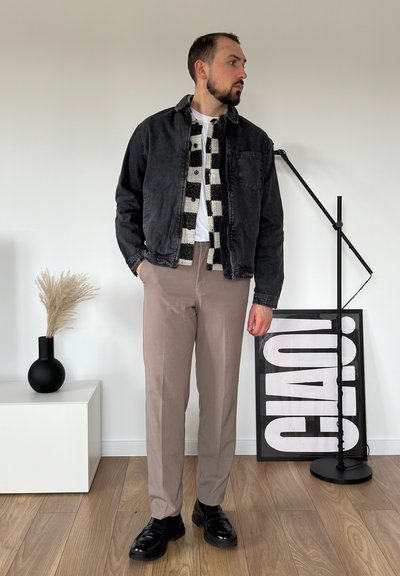 Chaqueta de mezclilla negra sobre una camisa blanca, combinada con una camisa de cuadros en blanco y negro y pantalones beige. Zapatos negros pulidos completan el look.