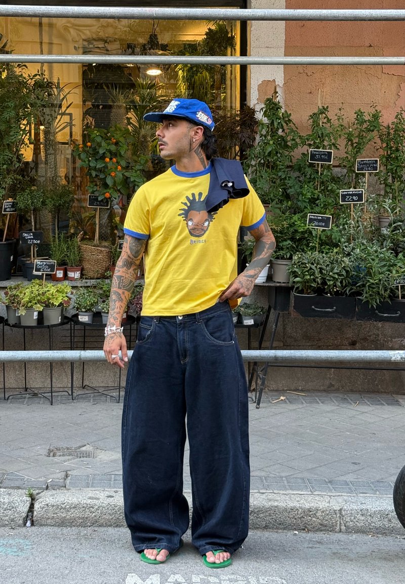 Homme avec des tatouages portant un t-shirt jaune graphique, une casquette bleue, un pantalon large foncé et des tongs vertes, se tient près d'une vitrine d'un magasin de plantes sur un trottoir.