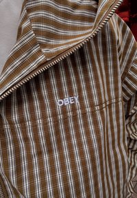 Hnědo-bílá pruhovaná zipová bunda s vyšitým logem "OBEY". Má lehkou tkaninu a vnitřní vrstvu se stejným vzorem.