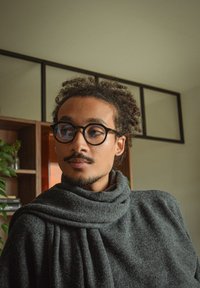 Runde svarte briller, grå strikket skjerf, teksturert ullstoff drapert, middels langt krøllete hår, mykt naturlig lys fra siden.